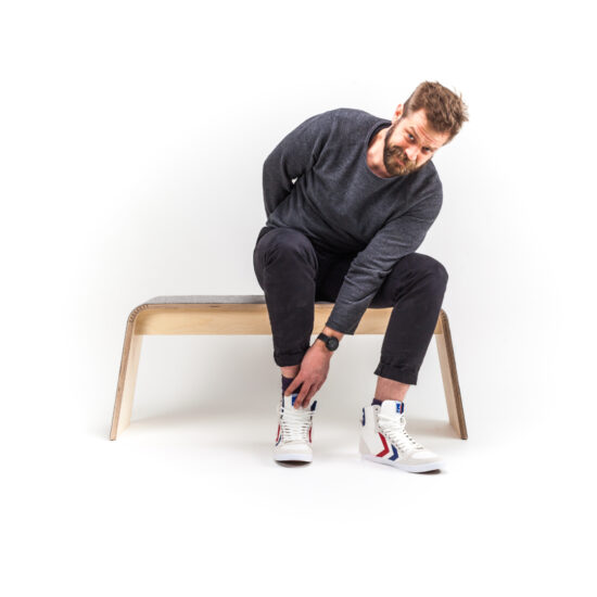 ein mann sitzt auf der sitzbank von stadig aus hellem holz und greift mit einer hand nach seinem schuh und schaut verschmitzt in die kamera